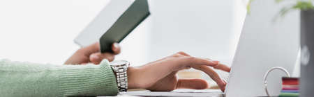 partial view of african american freelancer holding notebook and typing on laptop on blurred...