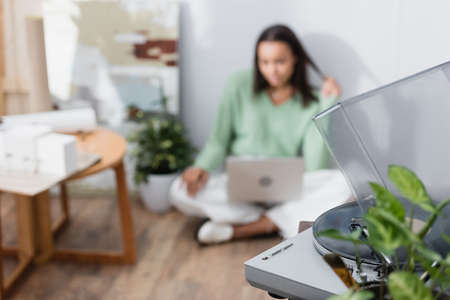 selective focus of record player and african american freelancer sitting on floor with laptop on...