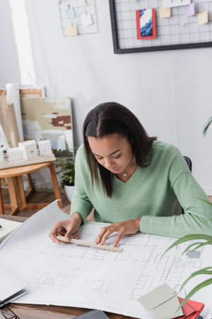 concentrated african american architect measuring blueprint with ruler at workplace
