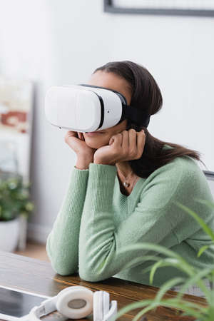 african american designer sitting in vr headset at workplace near headphones, blurred foreground