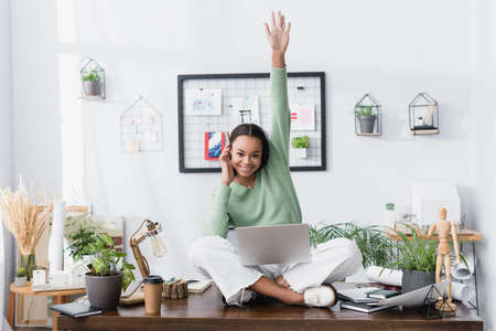 excited african american architect looking at camera while sitting on desk with laptop and raised handの写真素材