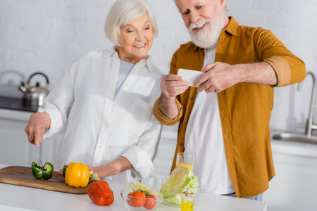 Fresh vegetables near smiling senior couple with smartphone cooking on blurred backgroundの写真素材