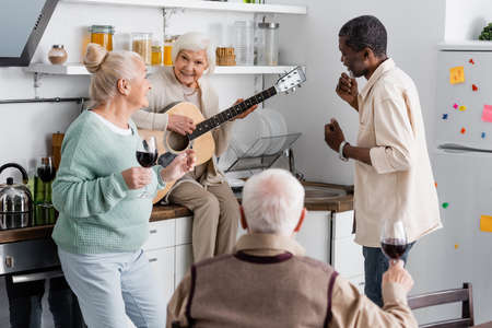 happy senior woman playing acoustic guitar near retired multicultural friends in kitchenの写真素材