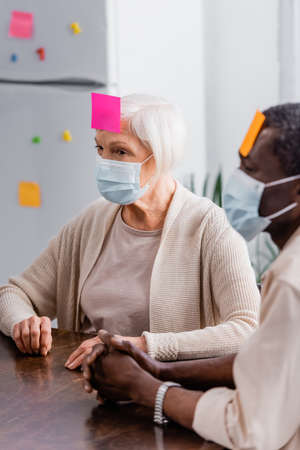 multicultural senior friends in medical masks with sticky notes on foreheads playing game in kitchenの写真素材