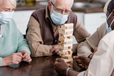 retired african american man in medical mask playing tower wood blocks game near friends on blurred backgroundの写真素材