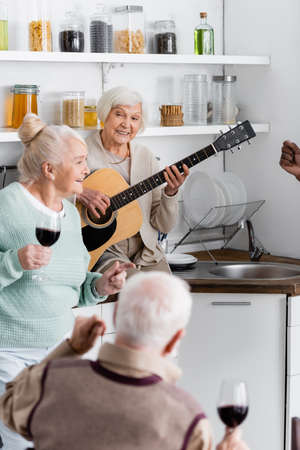 happy retired woman playing acoustic guitar near senior interracial friends in kitchenの写真素材