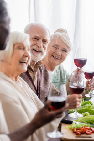 cheerful interracial pensioners laughing while holding glasses with wineの写真素材