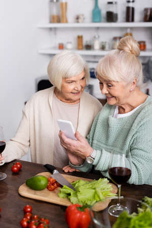 happy senior woman holding smartphone near retired friend in kitchenの写真素材