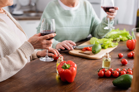 cropped view of senior women holding glasses of red wine near fresh vegetables in kitchenの写真素材