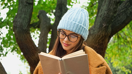 young woman in knitted beanie and eyeglasses reading novel in park