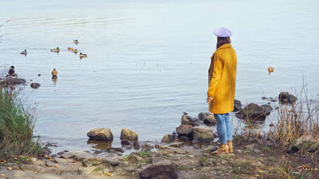 woman in stylish autumn outfit standing near lake with wild ducks in park