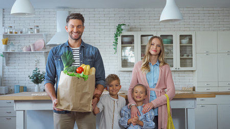 Smiling man holding paper bag with fresh food near family with kids in kitchen