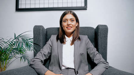 Smiling businesswoman sitting in armchair near plant in officeの写真素材
