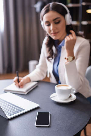 freelancer in headphones writing in notebook near laptop and smartphone with blank screen, blurred...