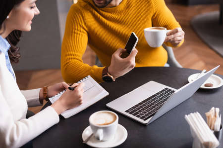 cropped view of freelancer showing smartphone to friend writing in notebook in restaurantの写真素材