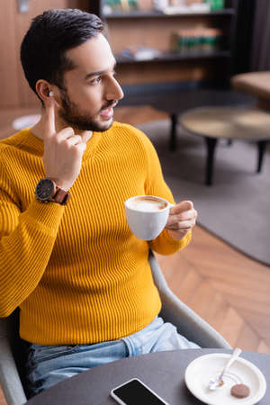 young arabian man looking away while adjusting earphone and holding cup of coffee in restaurantの写真素材