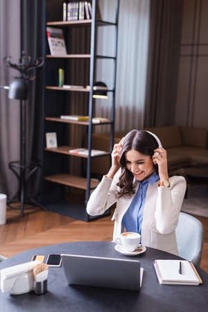 cheerful freelancer putting on headphones while sitting in restaurant near laptopの写真素材