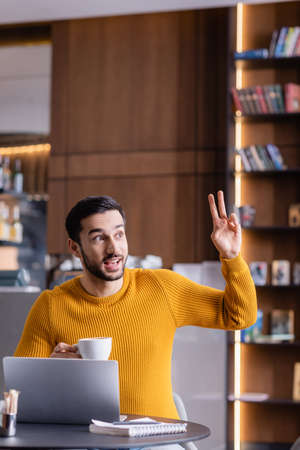happy arabian freelancer showing victory gesture while holding cup of coffee in restaurant