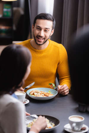 Cheerful arabian man holding cutlery near delicious salad, cappuccino and girlfriend on blurred foreground in restaurantの写真素材