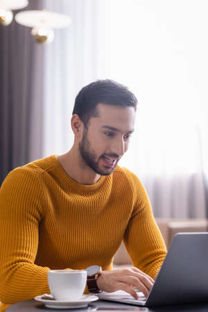 young arabian freelancer smiling while typing on laptop near cup of coffee in restaurantの写真素材