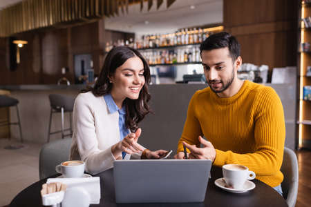 Multiethnic friends looking at laptop near coffee on blurred foreground in restaurant
