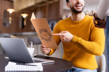partial view arabian teleworker pointing at menu card near waitress in restaurant, blurred...