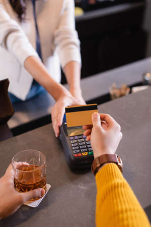Cropped view of man with whiskey paying with credit card near bartender with terminal on blurred...