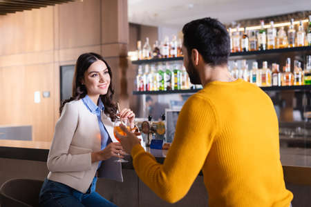 Smiling woman with cocktail looking at muslim friend on blurred foreground near bar counter in...