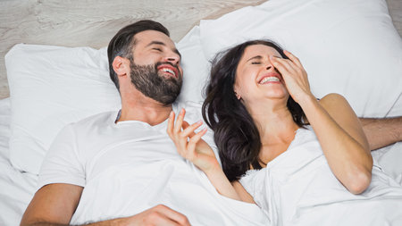 laughing woman touching face while lying in bed near happy man