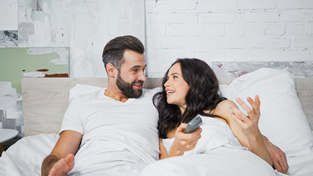 cheerful couple looking at each other while watching tv in bedの写真素材