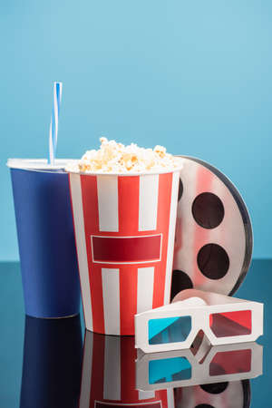 bucket with popcorn, cup with soda, film bobbin and 3d cinema glasses on glossy surface isolated on blueの写真素材