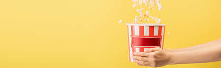 partial view of woman holding striped bucket of popcorn isolated on yellow, banner, cinema concept