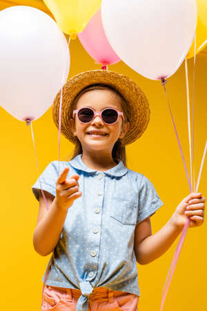 happy kid in straw hat and sunglasses holding balloons and pointing with finger isolated on yellow