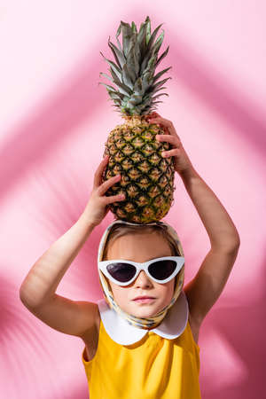 girl in headscarf and sunglasses holding pineapple above head on pinkの写真素材