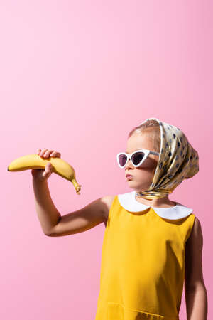 girl in headscarf and sunglasses puffing cheeks while holding banana isolated on pinkの写真素材