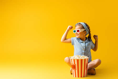 smiling kid in 3d glasses sitting with crossed legs near popcorn bucket on yellowの写真素材