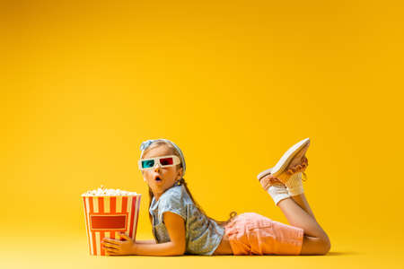 shocked girl in 3d glasses lying near popcorn bucket on yellowの写真素材