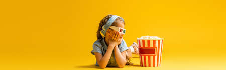 happy kid in 3d glasses lying near popcorn bucket on yellow, bannerの写真素材