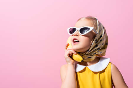 girl in headscarf and sunglasses holding banana near ear isolated on pinkの写真素材