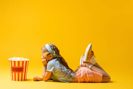 side view of kid in 3d glasses lying near popcorn bucket on yellowの写真素材
