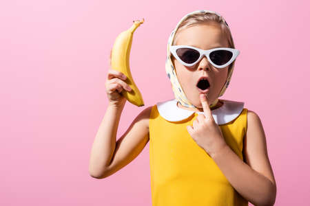 shocked girl in headscarf and sunglasses holding banana isolated on pinkの写真素材
