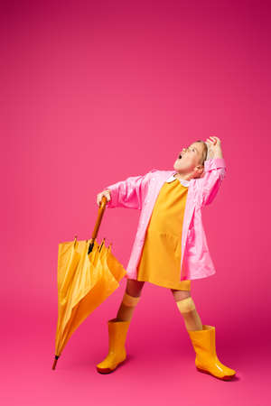 full length of surprised girl in raincoat and rubber boots standing with umbrella on crimsonの写真素材