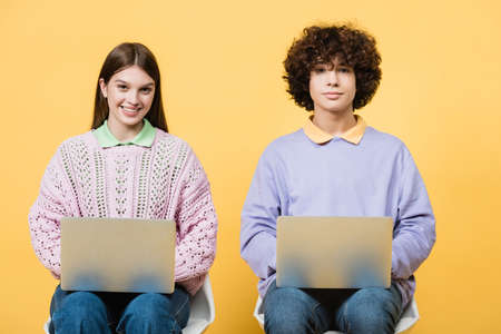 Teen girl with laptop looking at camera near friend isolated on yellowの写真素材