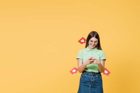 Smiling teenager with paper hearts on sticks using smartphone isolated on yellow