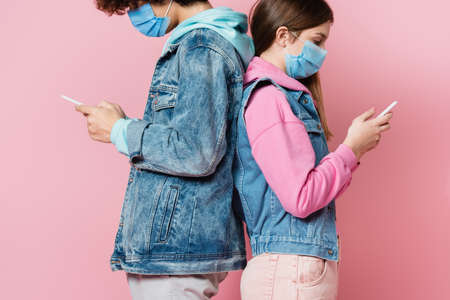 Side view of teenagers in medical masks using cellphones back to back isolated on pink