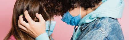 Curly teenager in medical mask touching face of girlfriend isolated on pink, banner
