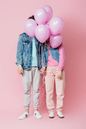 Teen couple holding hands while balloons covering faces on pink backgroundの写真素材