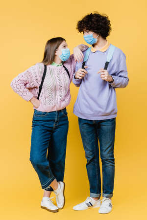 Girl in medical mask looking at curly friend on yellow background