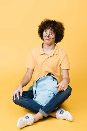 Confused curly boy in eyeglasses sitting near backpack on yellow background