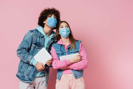 Teen boy and girl in medical masks holding digital tablets and looking away on pink backgroundの写真素材